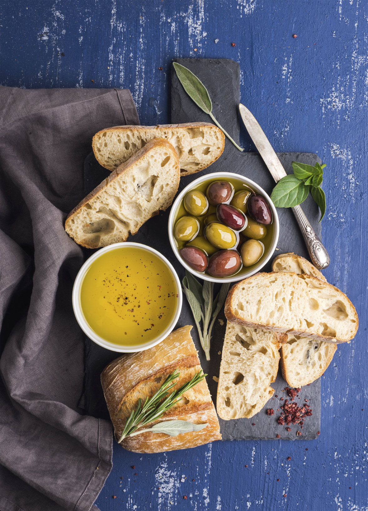 Mediterranean snacks set. Olives, oil, herbs and sliced ciabatta bread on black slate stone board over painted dark blue background, top view.
